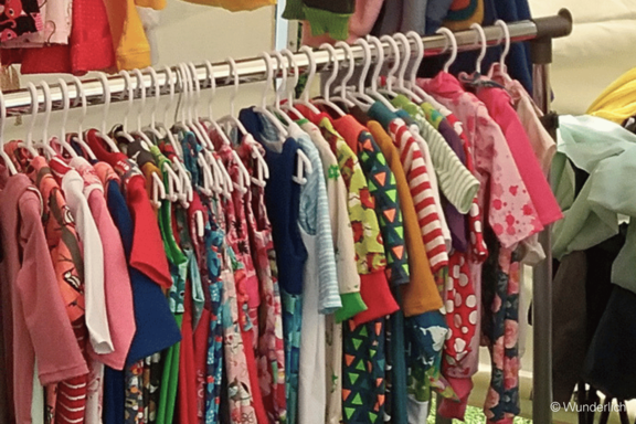 Bunte Kinderkleidung auf einer Stange: viele kleine Kleider und Shirts auf weißen Kleiderbügeln, in verschiedenen Farben und Mustern (Streifen, Blumen, Punkte) dicht nebeneinander aufgehängt.