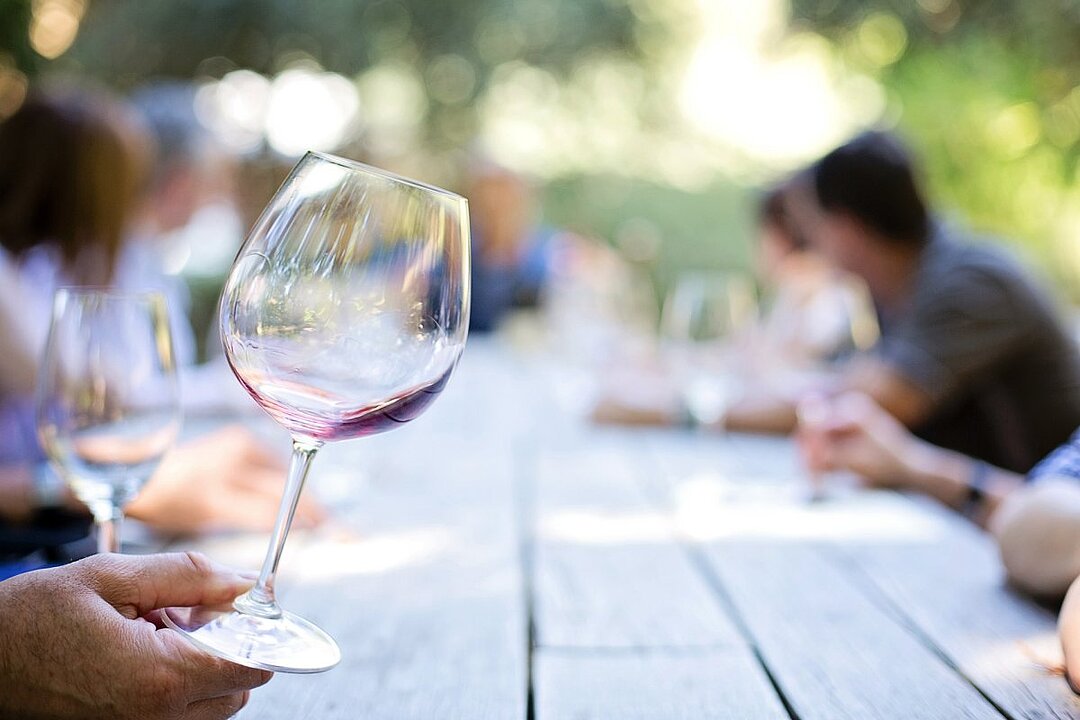 Eine Hand hält ein Glas Rotwein an einem gedeckten Tisch im Freien. (KI generiert)