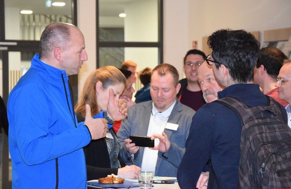 Mehrere Personen stehen um einen Stehtisch und unterhalten sich; ein Mann in blauer Jacke gestikuliert, andere hören zu, einer zeigt etwas auf einem Smartphone.