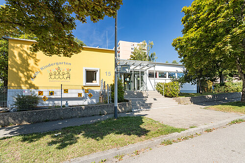 Vorderansicht eines gelben Kindergartengebäudes mit Kinderzeichnungen an der Fassade und Hausnummer 15, Glasüberdachung über dem Eingang, Treppen und Rampe, umgeben von Bäumen und Grünfläche.