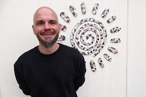 Ein lächelnder, glatzköpfiger Mann mit Bart in schwarzem Pullover steht vor einer weißen Wand, auf der ein Mosaik in Form einer spiraligen Sonne angebracht ist.