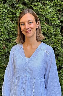 Lächelnde junge Frau mit schulterlangen braunen Haaren und hellblauer Bluse vor dichter grüner Hecke.