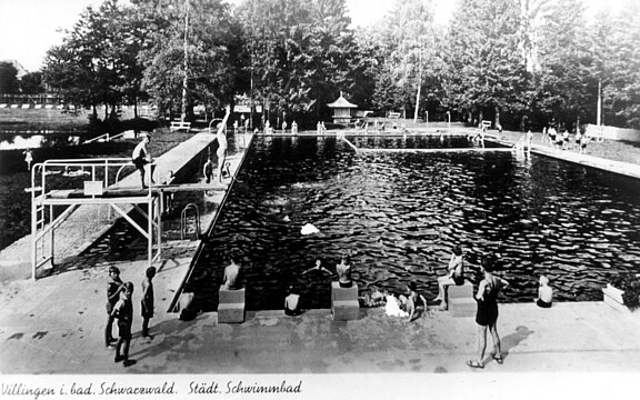 Schwarzweiß-Foto eines Freibads: großes rechteckiges Becken mit Sprungbrettern, mehreren Schwimmern und Personen am Beckenrand, umgeben von hohen Bäumen.
