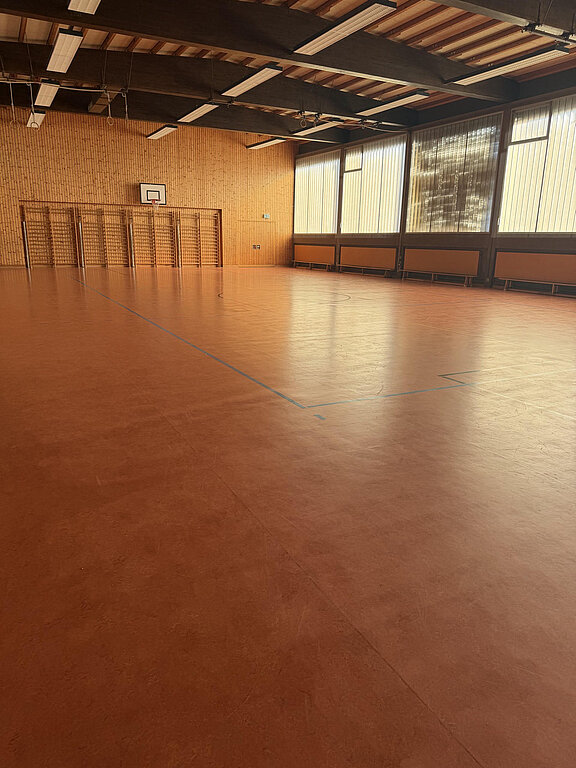 Leere Turnhalle mit braunem Hallenboden, Markierungen für Spielfeld, Basketballkorb an der Stirnseite und Sprossenwänden; große Fenster lassen Tageslicht herein.
