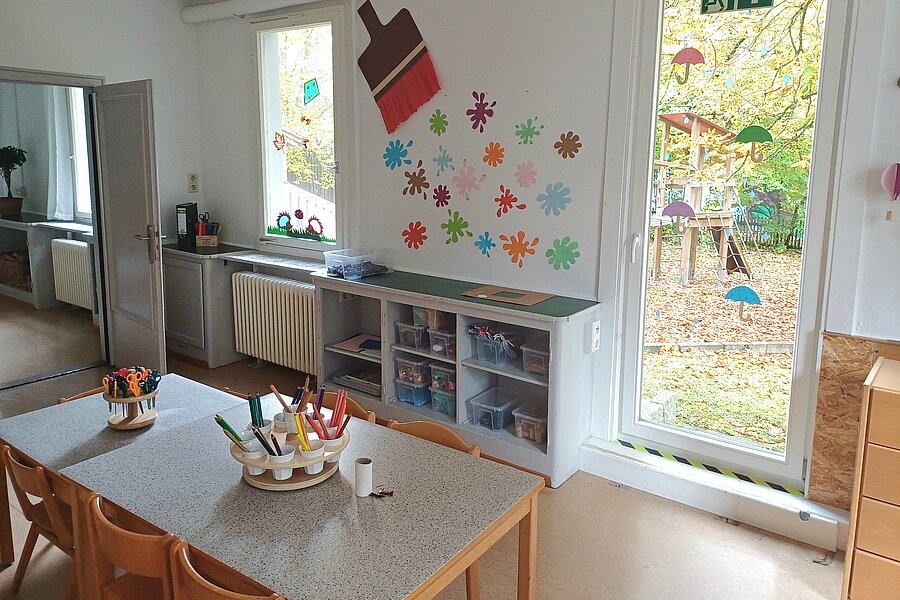 Heller Gruppenraum einer Kita mit Tisch und Stühlen, Bastelmaterial in Behältern, Regal mit Plastikkisten, bunter Wanddekoration (großer Pinsel und Blumen) und einer großen Glastür zum Außenspielplatz.