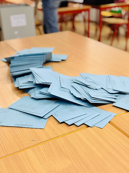 Gestapelte und verstreute blaue Wahlzettel liegen auf einem Holztisch, bereit zur Auszählung.