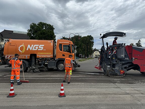 Feinfräsen Deckensanierung Bertholdstraße Zwei Baufahrzeuge, darunter eine große Fräsmaschine und eine Kehrmaschine, fahren entlang der Bertholdstraße und bearbeiten die Straße. Neben den Fahrern sind zwei Bauarbeite stehen im Vordergrund zu sehen. Einer von ihnen hält einen Besen.