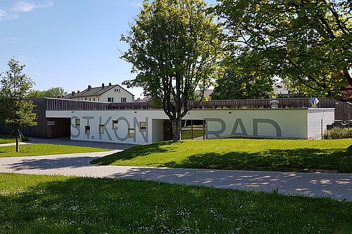 Niedriges weißes Gebäude mit großer beschrifteter Fassade, umgeben von Rasen, Fußweg und Bäumen an einem sonnigen Tag.