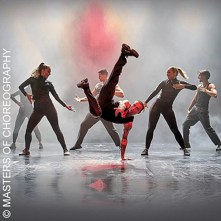 Fünf Tänzer in dynamischer Choreografie auf hellem Bühnenboden, einer in roter Hose. (KI generiert)