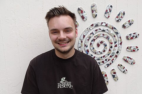 Junger lächelnder Mann mit kurzem dunklem Haar und schwarzem T‑Shirt steht vor weißer Wand, daneben große Mosaiksonne in Spiralform.