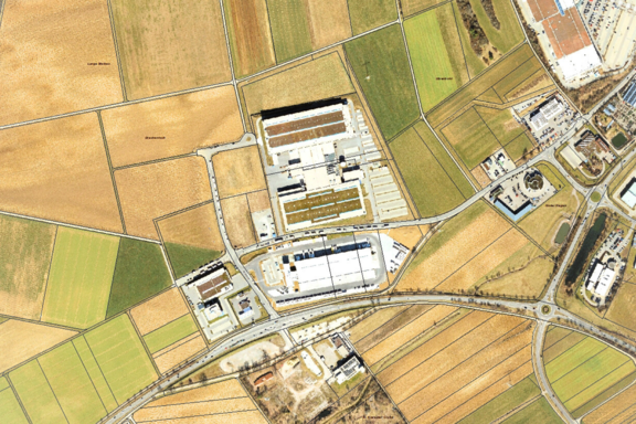 Luftaufnahme eines Industrie- und Logistikareals mit mehreren großen Hallen, Ladezonen und Parkflächen, umgeben von rechteckigen Ackerfeldern sowie angrenzenden Straßen und einem Kreisverkehr.