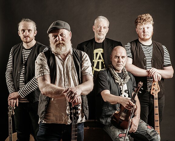 Fünf Männer posieren vor neutralem Hintergrund als Folk-/Rock-Band; sie halten akustische Gitarren, E‑Bass und Geige, tragen Westen und gestreifte Hemden und blicken ernst in die Kamera.