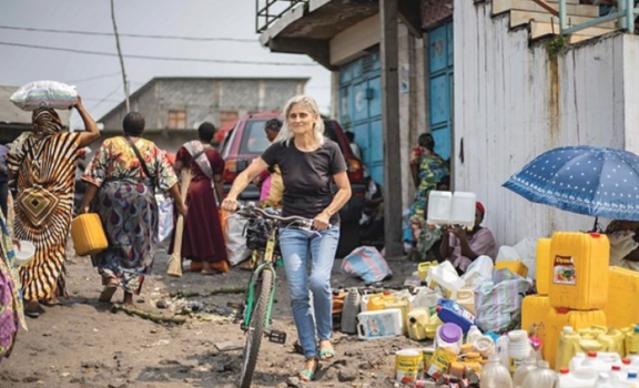 Blonde Frau schiebt ein Fahrrad durch einen belebten Straßenmarkt; um sie herum Frauen in bunten Kleidern tragen Waren, Verkäufer bieten Wasserkanister und Haushaltsbehälter an.