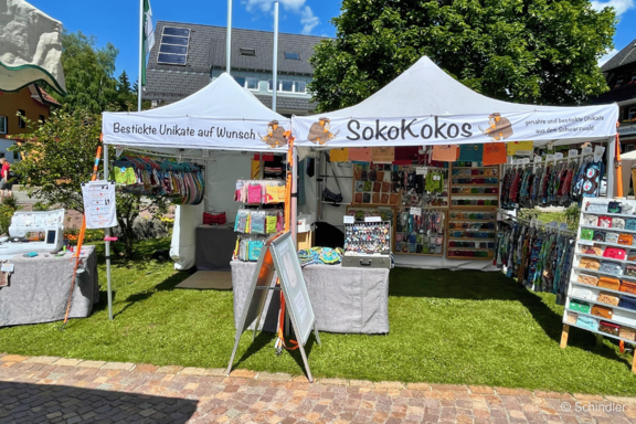 Marktstand mit zwei weißen Pavillons "SokoKokos", präsentiert bunte, handgefertigte Stoffwaren, Taschen, Accessoires und bestickte Unikate auf Regalen und Tischen.