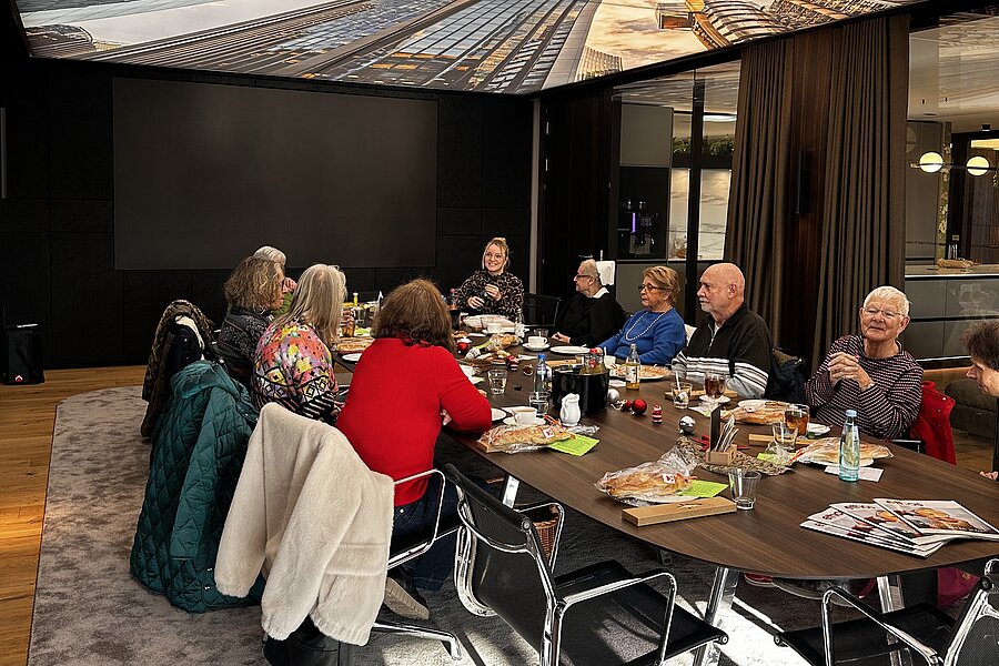 Ältere Menschen sitzen in einem modernen Konferenzraum um einen Tisch mit Getränken und Snacks, im Gespräch. An der Decke hängt ein großes Bild einer städtischen Skyline und des Himmels.