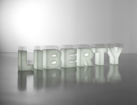 Große dreidimensionale Blockbuchstaben bilden das Wort "LIBERTY", auf jeder Oberseite Reihen von Metallspikes; die Buchstaben stehen auf spiegelnder Oberfläche.