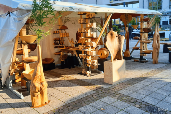 Marktstand mit weißem Pavillon, darin handgefertigte Holzskulpturen, Schalen und Möbelstücke auf Regalen und Podesten, warme Holzfarben und organische Formen, arrangiert als Verkaufsausstellung auf einem städtischen Platz.