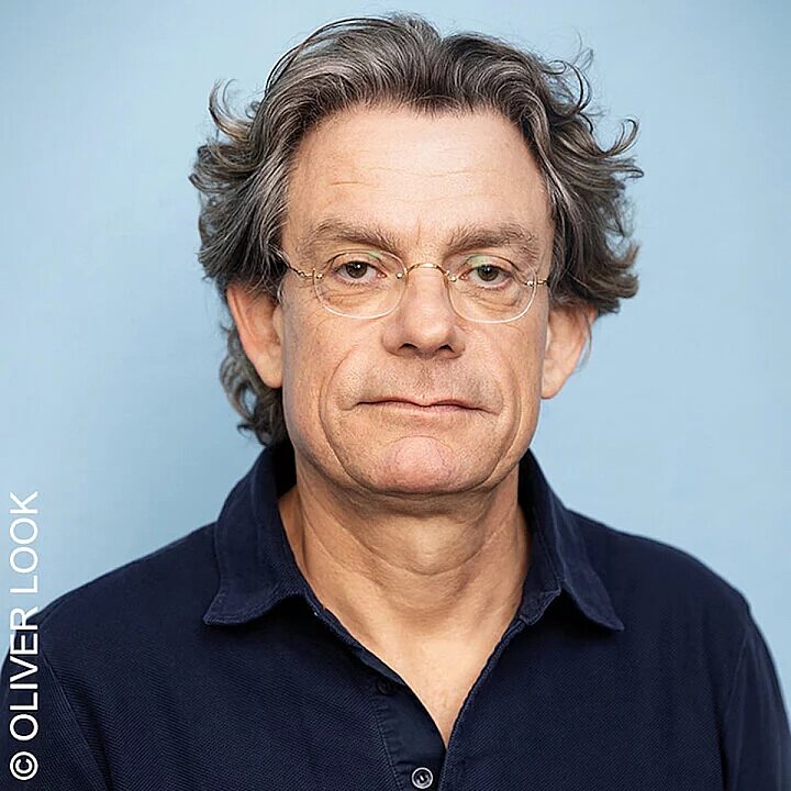 Porträt eines lächelnden älteren Mannes mit Brille und lockigem Haar in dunklem Pullover. (KI generiert)