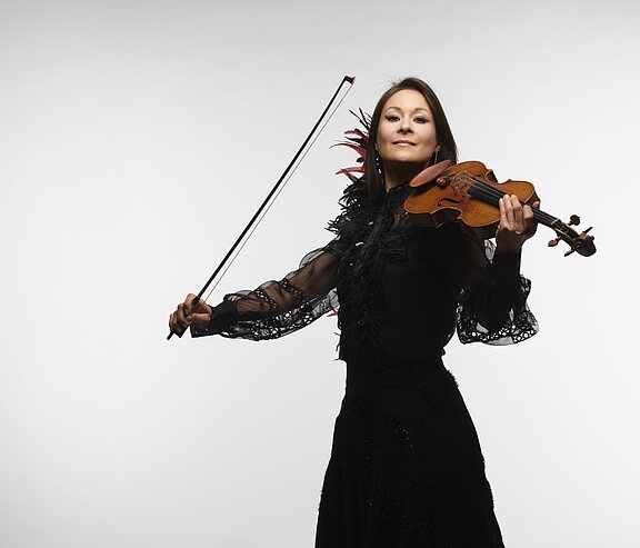 Stehende Violinistin in schwarzem Kleid mit erhobenem Bogen, spielt Geige und blickt selbstbewusst in die Kamera.