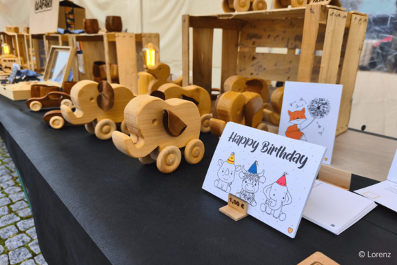 Marktstand mit handgefertigten Holzspielzeugen: mehrere Elefanten auf Rädern und Holzautos auf einem Tisch, im Vordergrund eine "Happy Birthday"-Karte mit gezeichneten Elefanten.