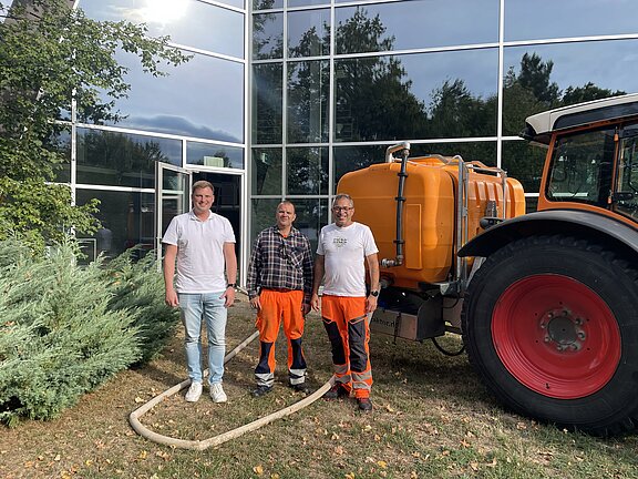 Drei Männer stehen neben einem orangen Tankanhänger, der an einen Traktor angehängt ist; zwei tragen orange Arbeitskleidung, einer Freizeitkleidung, ein Schlauch liegt auf dem Rasen vor einem Gebäude mit großen Glasfenstern.