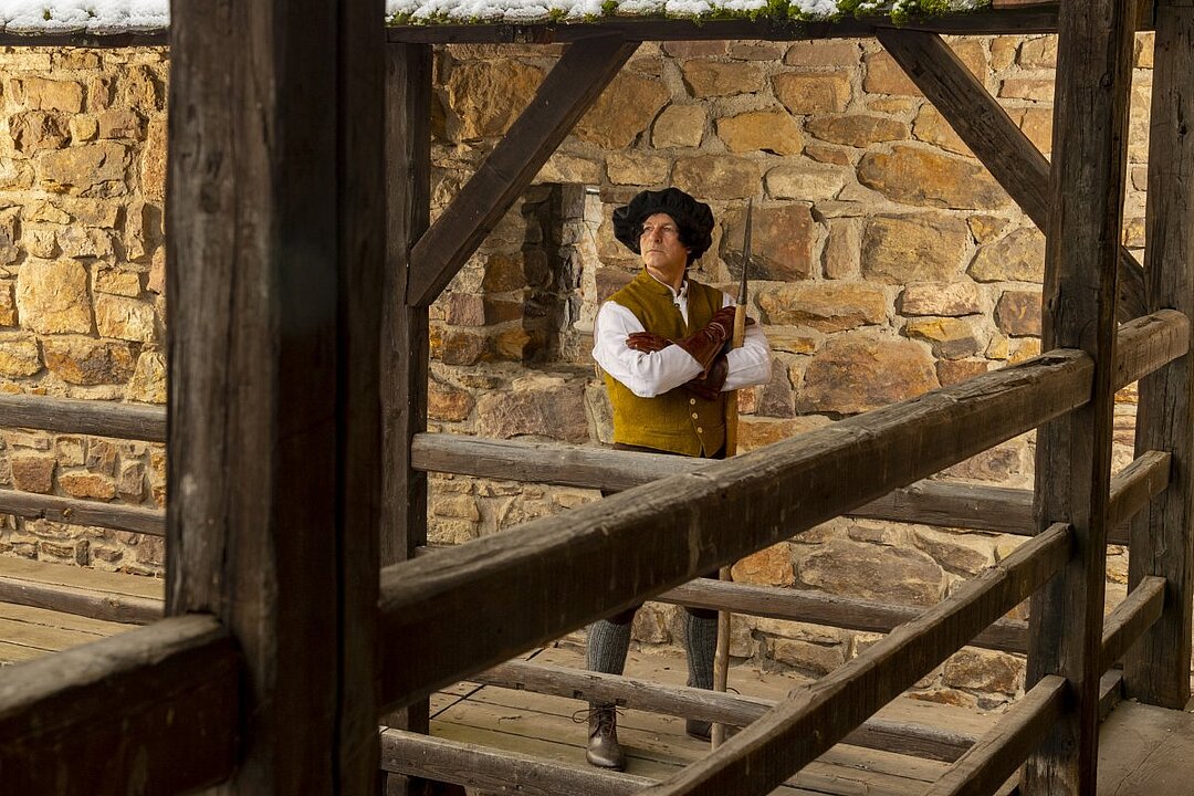 Ein Mann in historischer Kleidung steht auf einem Holzpodest vor einer Steinmauer. (KI generiert)