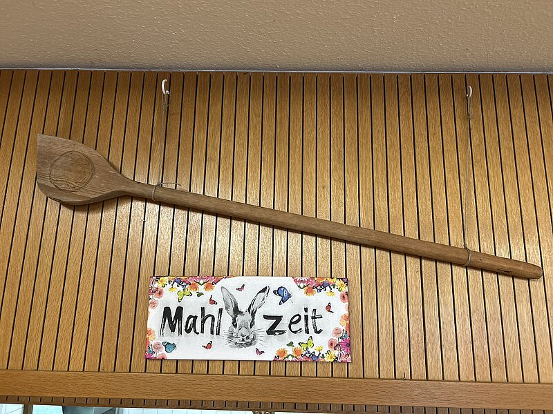 Großer hölzerner Kochlöffel an einer Holzvertäfelung, darunter ein buntes Schild mit der Aufschrift „Mahlzeit“ und einer Hasenzeichnung.