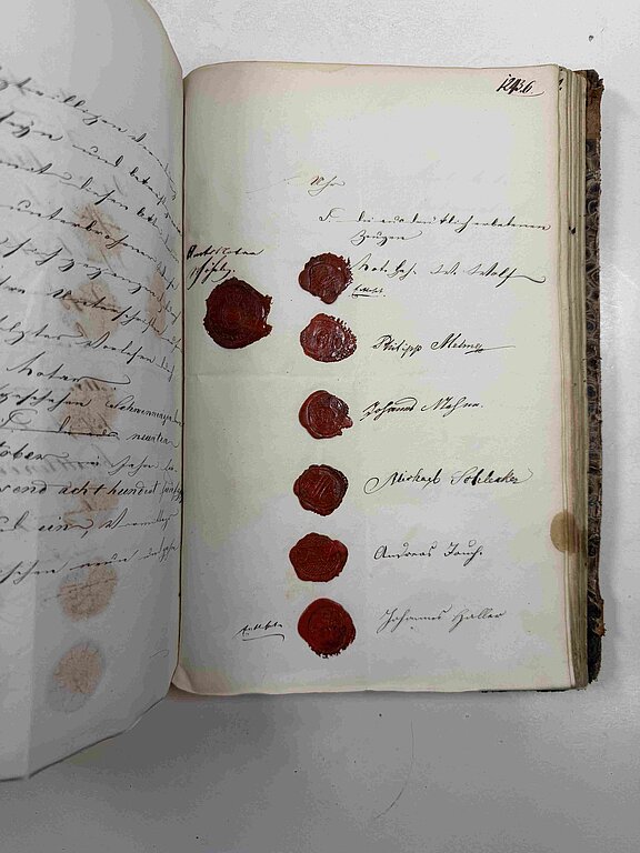 Geöffnetes altes Buch mit handschriftlichen Einträgen und einer senkrechten Reihe von sieben roten Wachssiegeln, jeweils neben Unterschriften und Namen.