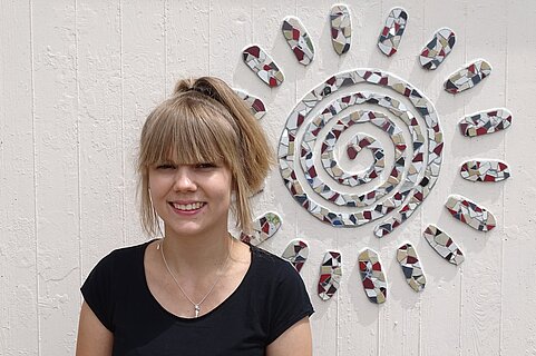 Lächelnde Person mit hellem Pferdeschwanz und schwarzem T‑Shirt steht vor weißer Holzwand, daneben großes Mosaik in Form einer spiralförmigen Sonne.