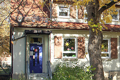 Fassadenansicht eines zweistöckigen hellen Hauses oder Bürogebäudes mit blauem Eingang, mehreren nebeneinanderliegenden Fenstern und herbstlich gefärbten Bäumen im Vordergrund.