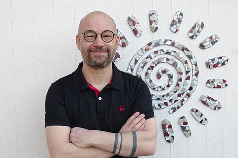 Lächelnder, glatzköpfiger Mann mit Brille und kurzem Bart, Arme verschränkt, vor weißer Wand mit spiraliger Mosaik‑Sonne aus bunten Fliesen.