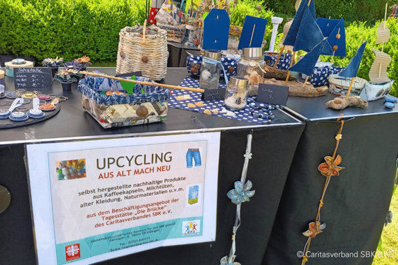 Marktstand mit Upcycling-Produkten: Tisch ausgestellt mit kleinen Segelbooten, Vogelhäuschen, Deko-Gläsern und handgefertigten Accessoires; Schild "Upcycling – Aus alt mach neu" weist auf nachhaltige, aus Altmaterialien hergestellte Waren hin.