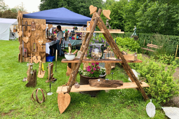 Marktstand im Freien mit rustikalem Holzdisplay: dreieckiges Regal mit Holzherzen, Dekorationsstücken, Glasgefäß und Topfpflanze.