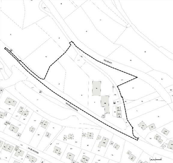 Schwarz-weißer Lageplan mit gestrichelter Begrenzung, die ein längliches Areal mit mehreren Gebäuden entlang einer Straße umschließt; umliegende Parzellen und einzelne Wohnhäuser sind schematisch eingezeichnet.