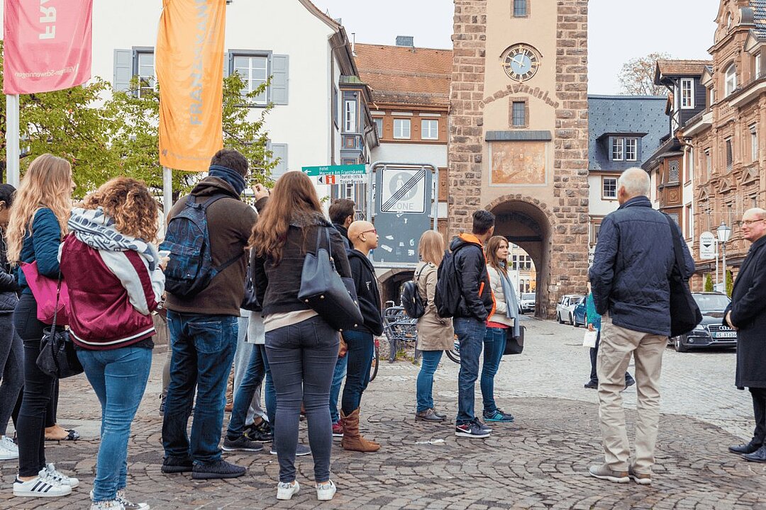 Gruppe von Menschen steht auf Kopfsteinpflaster vor historischem Tor mit Uhr und Fahnen. (KI generiert)