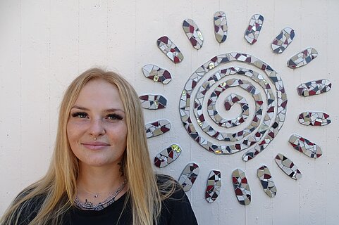 Frau mit langen blonden Haaren lächelt vor einer weißen Wand neben einer Mosaikspirale, die eine stilisierte Sonne bildet.