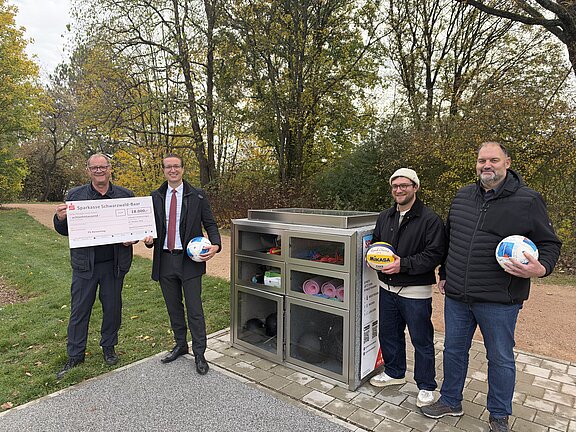 Vier Männer stehen neben einem Sportgeräteautomaten im Freien, einer hält einen großen Scheck über 18.000 Euro.