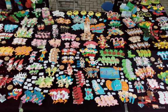 Marktstand mit schwarzem Tischtuch, bedeckt von zahlreichen bunten handgefertigten Holzfiguren, Magneten und Anhängern in Form von Tieren, Blumen und Früchten, ordentlich in Reihen angeordnet.
