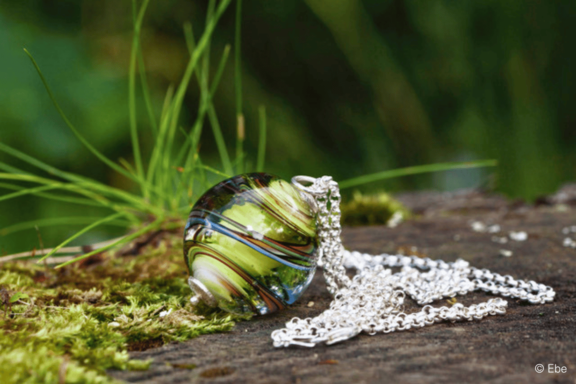 Nahaufnahme eines runden Glasanhängers mit grün-blauem Wirbelmuster an einer silbernen Kette, auf Moos und Holz liegend.