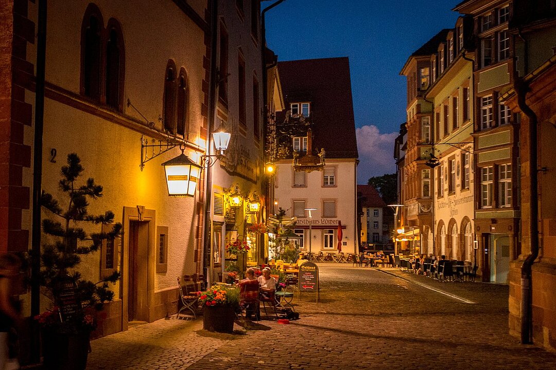 Abendliche Gasse mit beleuchteten Gebäuden, Tischen und Stühlen vor einem Restaurant, zwei Personen sitzen an einem Tisch. (KI generiert)