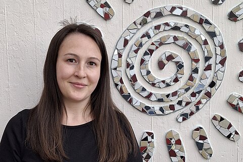 Porträt einer lächelnden Frau mit dunklen langen Haaren in schwarzem Shirt, stehend links neben einer bunten Mosaikspirale an der Wand.