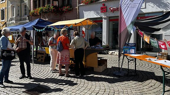 Menschen stehen an bunten Infoständen auf einem gepflasterten Marktplatz, betrachten Informationsmaterial und unterhalten sich.