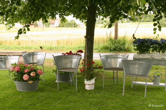 Sitzbereich im Garten unter einem Baum: mehrere verzinkte Metallstühle und -tische, blühende Blumen in Metallkübeln, Feld im Hintergrund.