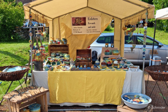 Marktstand unter gelbem Zelt mit zahlreichen bunten, handgefertigten Keramikschalen, Tassen und Vasen auf einem Tisch ausgestellt.