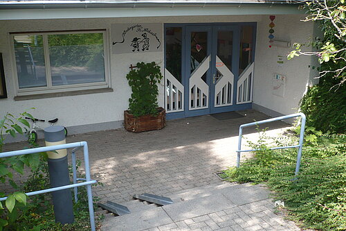 Eingangsbereich eines Hauses: blaue Doppeltür mit Fenstern, Treppe mit Handläufen und kleiner Rampe, Pflanzkübel und Wandbild mit Kindermotiv.
