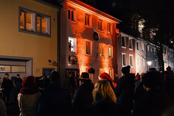 Abendliche Menschenmenge vor einem orange beleuchteten Haus mit großem Weihnachtskranz an der Fassade; Bewohner schauen aus geöffneten Fenstern, festliche Atmosphäre bei einer Weihnachtsveranstaltung.