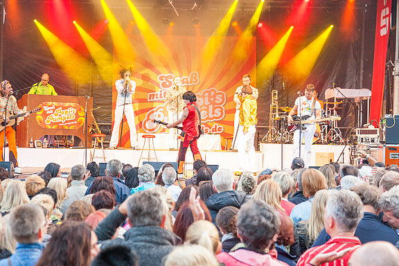 Band-Auftritt auf einer Bühne vor großem Publikum: Leadsänger vorn, Musiker mit Gitarre, Bläser und Schlagzeug, bunte Scheinwerfer und begeisterte Zuschauer.