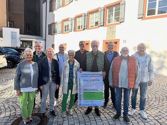 Zehn ältere Personen stehen auf einem gepflasterten Hof vor einem hellen Haus; zwei halten ein Plakat für einen städtischen Seniorentag.