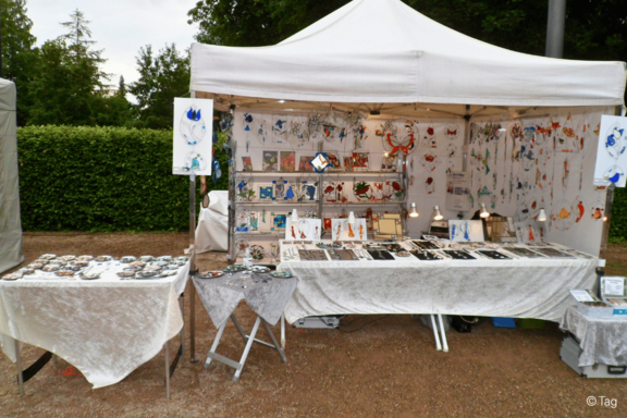 Marktstand mit weißem Zelt, Tischen und Regalen, auf denen handgefertigter Schmuck, Anhänger und kleine Kunstdrucke ausgestellt sind; bunte Dekorationen an der Rückwand.