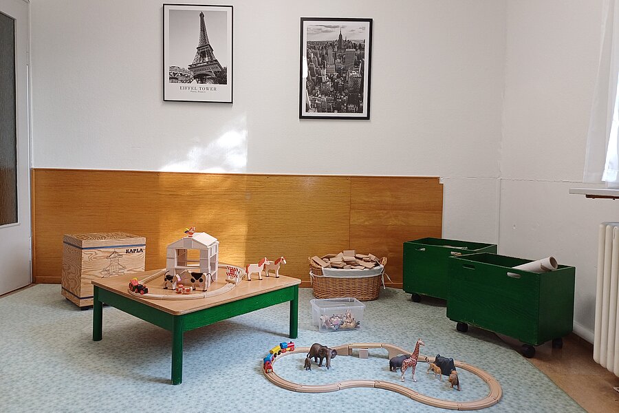 Heller Kinderspielraum mit niedrigem Tisch, Holzbauernhof, Holztiere und auf dem Boden geschlossener Holzeisenbahnkreis; grüne Spielkisten und zwei Schwarzweiß-Fotografien an der Wand.
